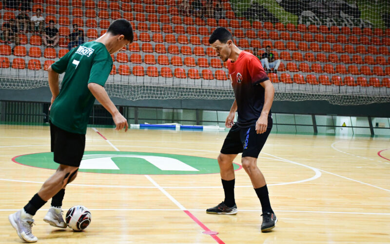 Dois jovens atletas jogam futsal em quadra coberta, disputando a bola com os pés em uma jogada próxima, enquanto mantêm postura de concentração, equilíbrio corporal e atenção aos movimentos do adversário durante a partida.