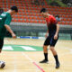 Dois jovens atletas jogam futsal em quadra coberta, disputando a bola com os pés em uma jogada próxima, enquanto mantêm postura de concentração, equilíbrio corporal e atenção aos movimentos do adversário durante a partida.