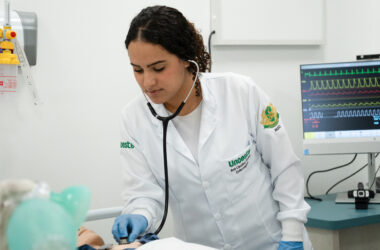 Estudante de Enfermagem da Unoeste realizando atendimento em laboratório, utilizando estetoscópio em manequim hospitalar enquanto monitora sinais vitais em equipamento médico, simulando prática clínica durante a graduação.