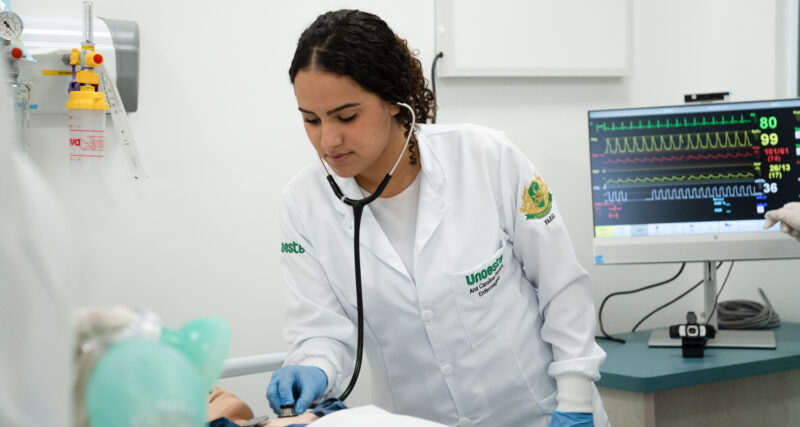 Estudante de Enfermagem da Unoeste realizando atendimento em laboratório, utilizando estetoscópio em manequim hospitalar enquanto monitora sinais vitais em equipamento médico, simulando prática clínica durante a graduação.