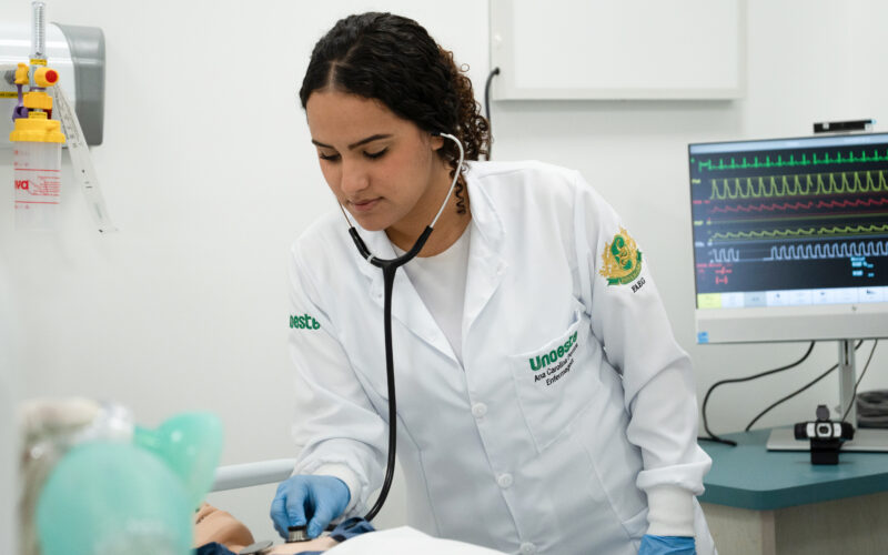 Estudante de Enfermagem da Unoeste realizando atendimento em laboratório, utilizando estetoscópio em manequim hospitalar enquanto monitora sinais vitais em equipamento médico, simulando prática clínica durante a graduação.