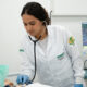 Estudante de Enfermagem da Unoeste realizando atendimento em laboratório, utilizando estetoscópio em manequim hospitalar enquanto monitora sinais vitais em equipamento médico, simulando prática clínica durante a graduação.