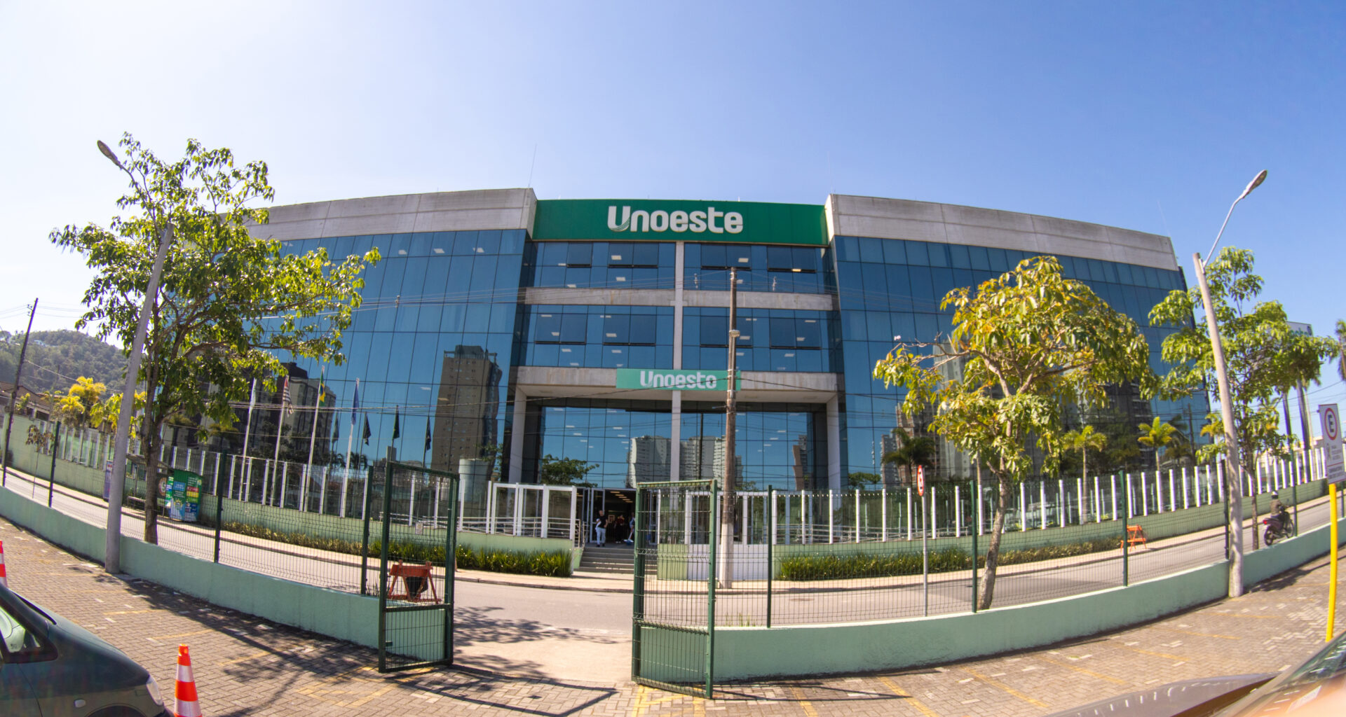 Fachada do campus da Unoeste no Guarujá, com prédio moderno de vidro, entrada principal e árvores ao redor.