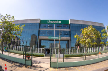 Fachada do campus da Unoeste no Guarujá, com prédio moderno de vidro, entrada principal e árvores ao redor.