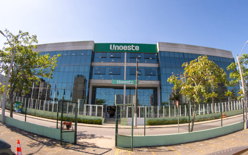 Fachada do campus da Unoeste no Guarujá, com prédio moderno de vidro, entrada principal e árvores ao redor.
