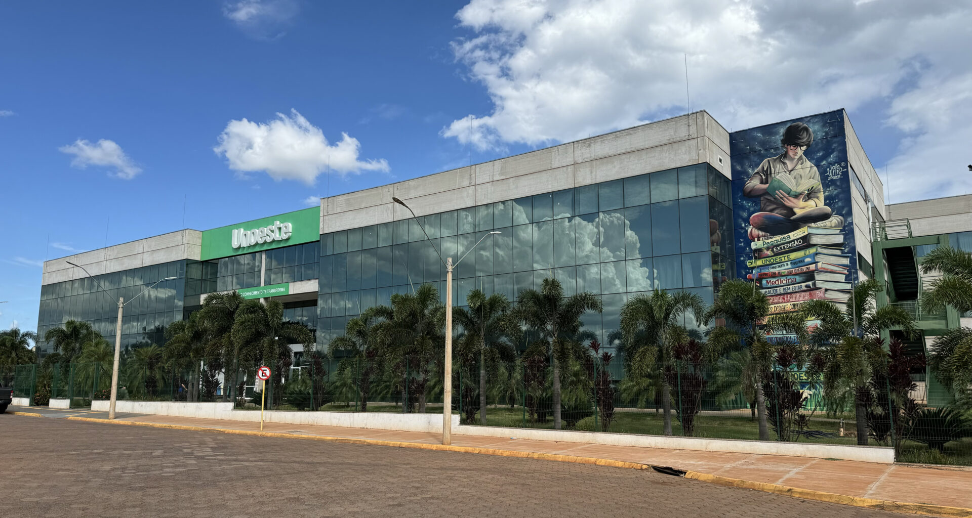 Fachada do campus da Unoeste em Jaú, com prédio moderno de vidro, área arborizada e mural artístico na lateral, sob céu azul com nuvens.