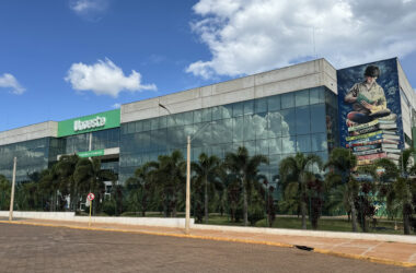 Fachada do campus da Unoeste em Jaú, com prédio moderno de vidro, área arborizada e mural artístico na lateral, sob céu azul com nuvens.