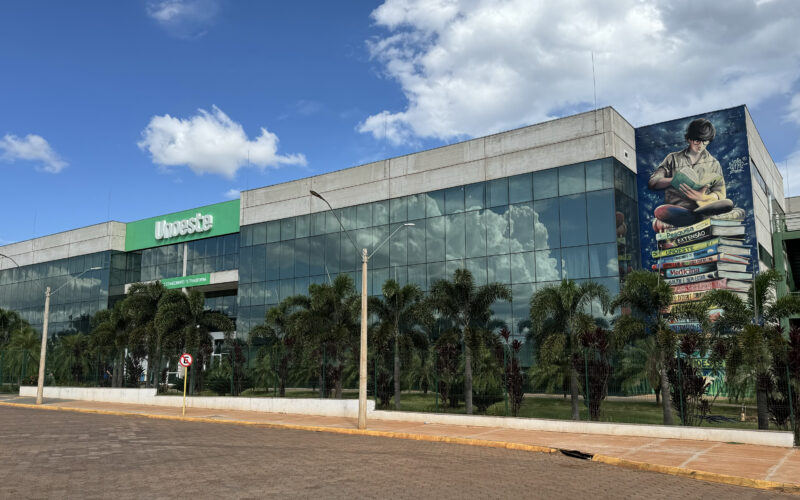 Fachada do campus da Unoeste em Jaú, com prédio moderno de vidro, área arborizada e mural artístico na lateral, sob céu azul com nuvens.