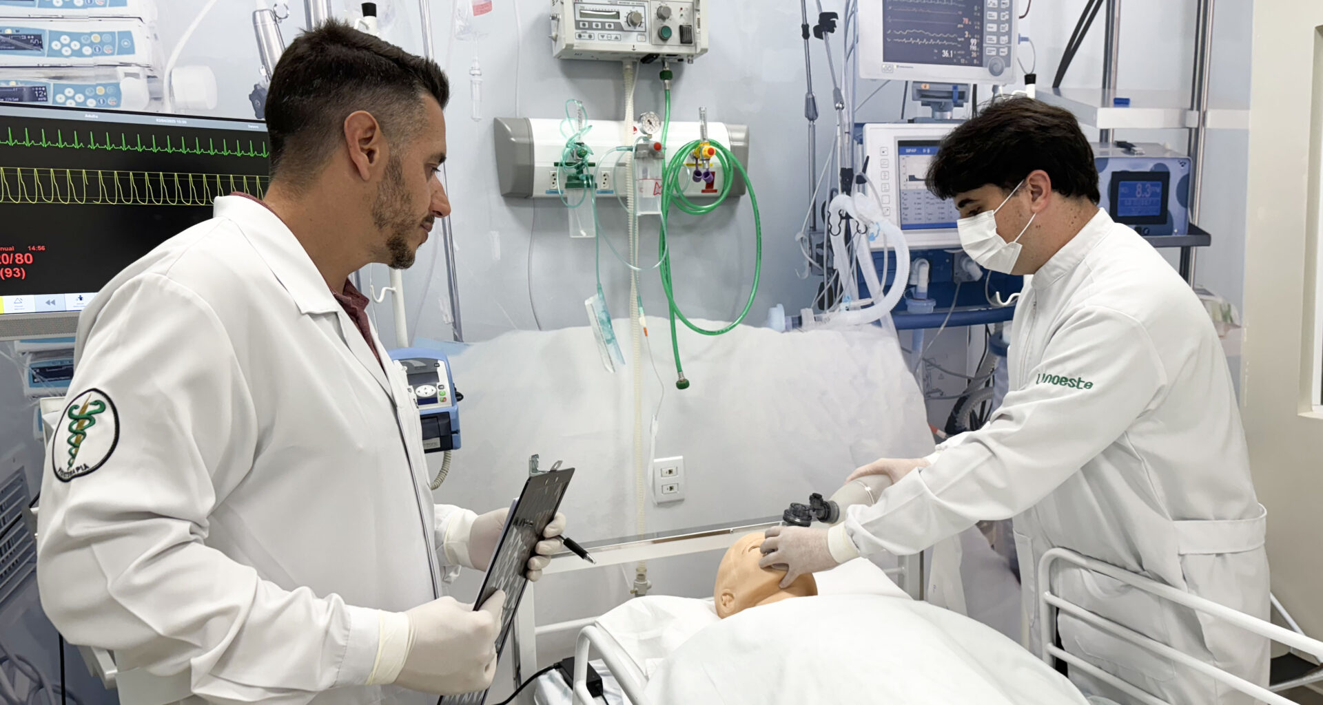 Fisioterapeutas em unidade de terapia intensiva realizam manejo de vias aéreas em paciente simulado, utilizando equipamentos hospitalares como ventilador mecânico e monitor multiparamétrico, ilustrando a prática profissional na UTI.