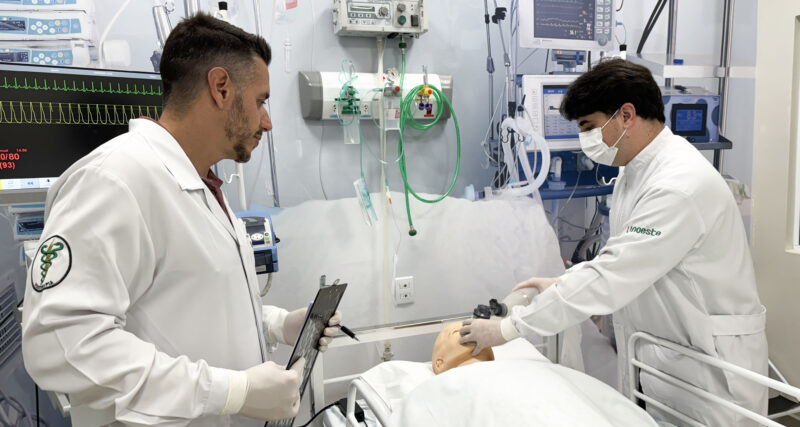 Fisioterapeutas em unidade de terapia intensiva realizam manejo de vias aéreas em paciente simulado, utilizando equipamentos hospitalares como ventilador mecânico e monitor multiparamétrico, ilustrando a prática profissional na UTI.