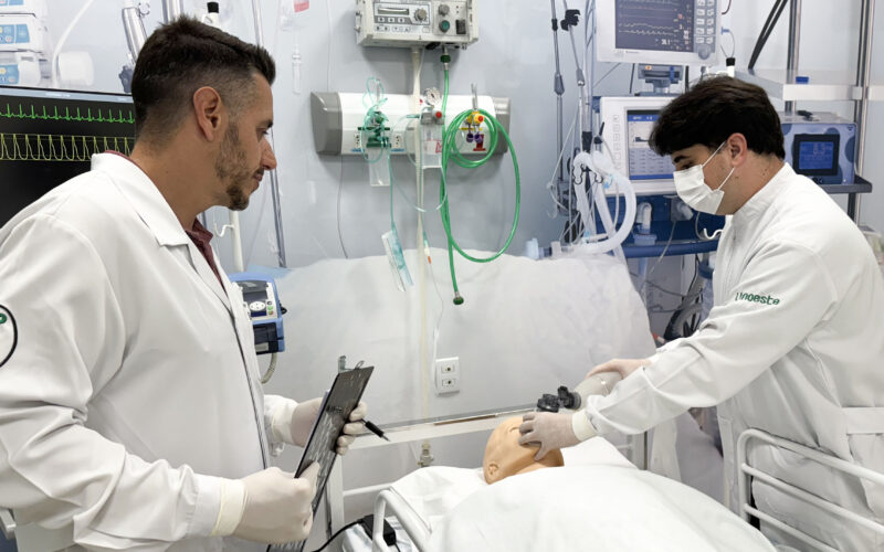 Fisioterapeutas em unidade de terapia intensiva realizam manejo de vias aéreas em paciente simulado, utilizando equipamentos hospitalares como ventilador mecânico e monitor multiparamétrico, ilustrando a prática profissional na UTI.