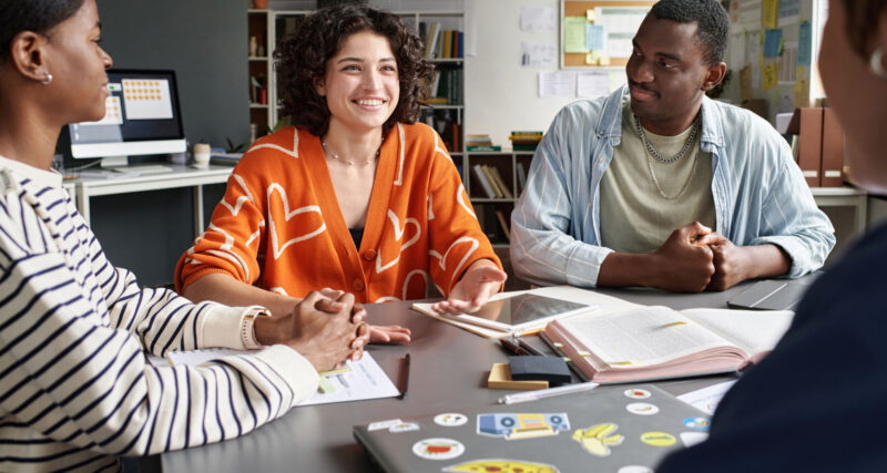 Grupo de jovens reunidos em ambiente de estudo ou trabalho colaborativo, sentados ao redor de uma mesa com notebook, tablet, livros e materiais de anotação. Uma jovem ao centro conversa e gesticula enquanto os demais escutam, em um cenário que representa troca de ideias, desenvolvimento de habilidades e preparação para o mercado de trabalho.