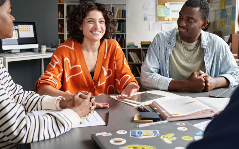 Grupo de jovens reunidos em ambiente de estudo ou trabalho colaborativo, sentados ao redor de uma mesa com notebook, tablet, livros e materiais de anotação. Uma jovem ao centro conversa e gesticula enquanto os demais escutam, em um cenário que representa troca de ideias, desenvolvimento de habilidades e preparação para o mercado de trabalho.