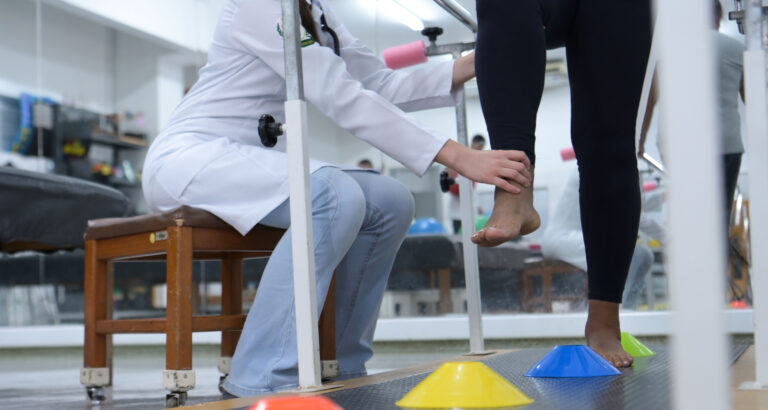 Pessoa em sessão de fisioterapia realizando exercício de equilíbrio e reabilitação dos membros inferiores, com acompanhamento de profissional de saúde em ambiente clínico equipado.