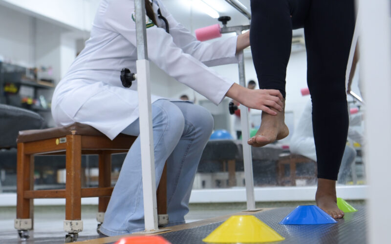 Pessoa em sessão de fisioterapia realizando exercício de equilíbrio e reabilitação dos membros inferiores, com acompanhamento de profissional de saúde em ambiente clínico equipado.