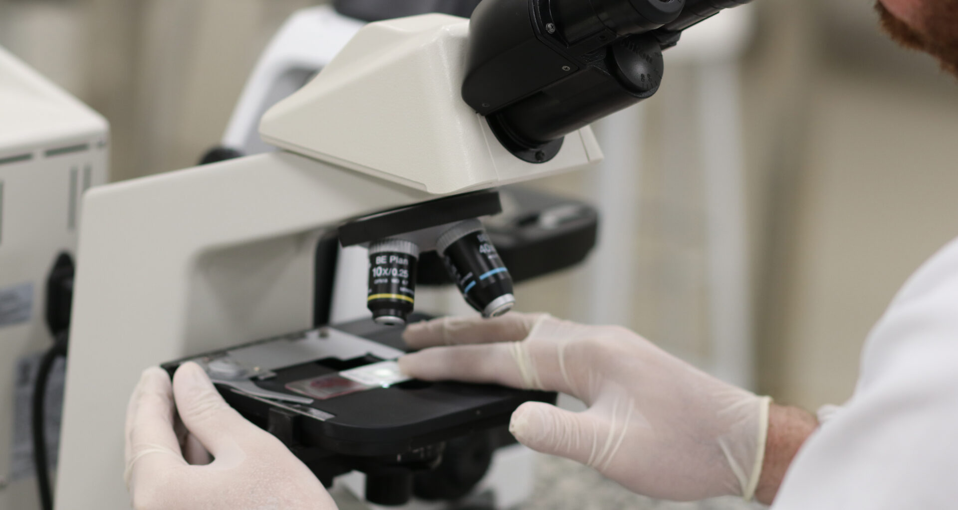 Pessoa utilizando microscópio em laboratório, com luvas, analisando lâmina com amostra para exame de citologia.