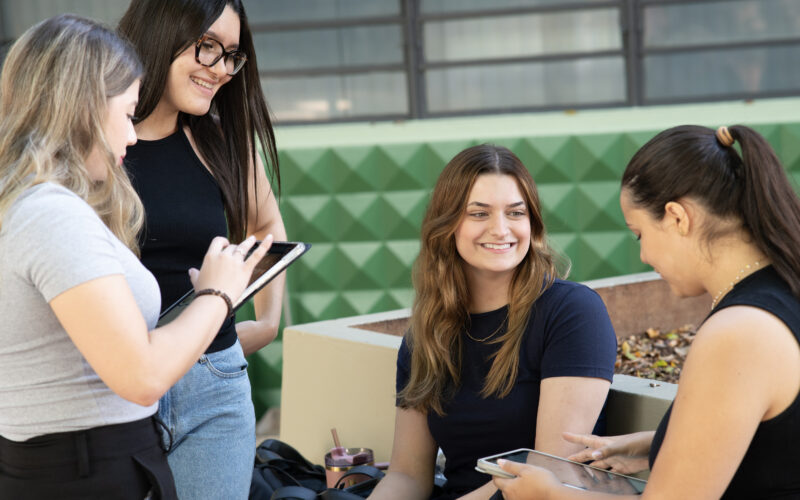 Grupo de estudantes conversando e estudando ao ar livre no campus universitário, utilizando tablets e materiais digitais durante a preparação para o Enem.