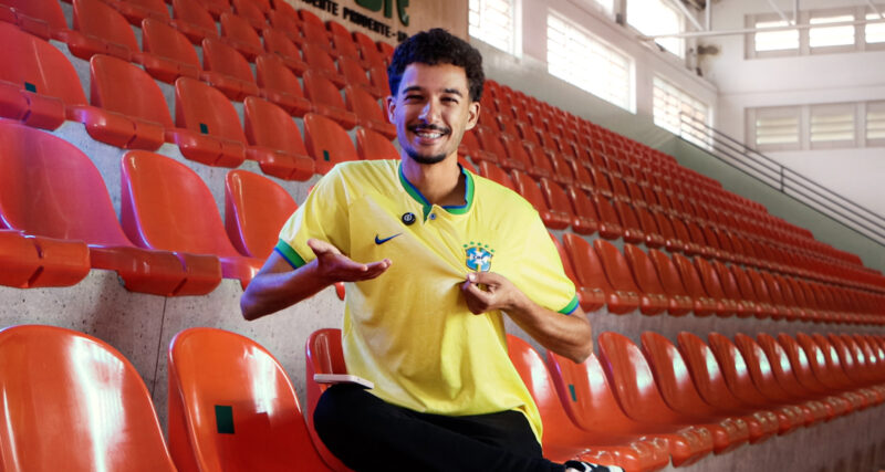 Homem sentado em arquibancada e usando camisa da seleção brasileira masculina de futebol.