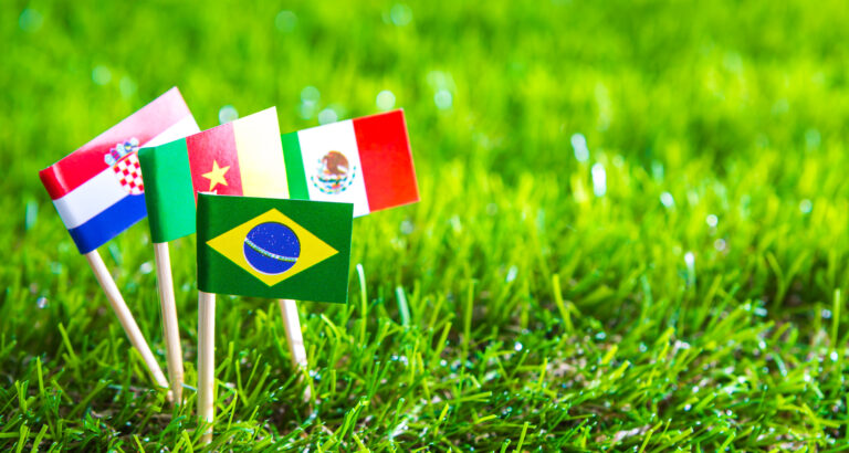 Bandeiras de diferentes países posicionadas sobre gramado de futebol, representando o clima internacional e multicultural da Copa do Mundo.
