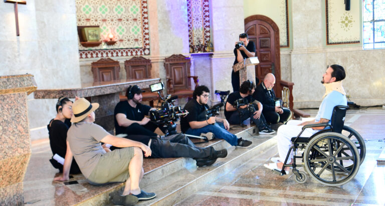 Equipe de filmagem posicionada dentro de uma capela, com câmeras, iluminação e operadores registrando uma cena enquanto um ator em cadeira de rodas, com colar cervical, atua à frente, em ambiente com altar, bancos de madeira e elementos religiosos ao fundo.
