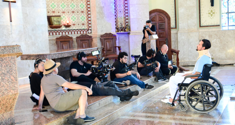 Equipe de filmagem posicionada dentro de uma capela, com câmeras, iluminação e operadores registrando uma cena enquanto um ator em cadeira de rodas, com colar cervical, atua à frente, em ambiente com altar, bancos de madeira e elementos religiosos ao fundo.