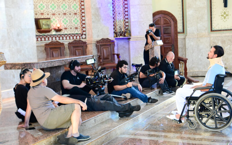 Equipe de filmagem posicionada dentro de uma capela, com câmeras, iluminação e operadores registrando uma cena enquanto um ator em cadeira de rodas, com colar cervical, atua à frente, em ambiente com altar, bancos de madeira e elementos religiosos ao fundo.