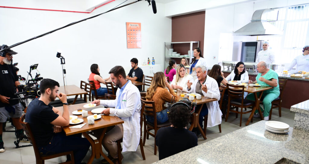 Equipe de filmagem registrando cena em um ambiente de cozinha/refeitório, com câmeras, microfone suspenso e iluminação posicionados, enquanto atores e figurantes sentados às mesas simulam uma refeição, incluindo profissionais caracterizados como médicos e equipe de saúde interagindo durante a gravação.