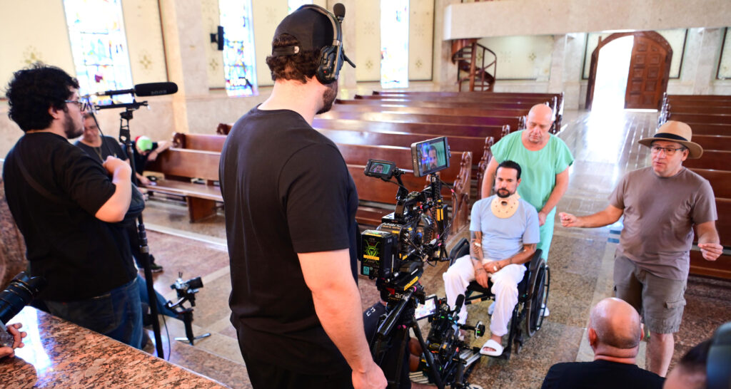 Equipe de filmagem dentro de uma capela, com câmera profissional, microfone e monitores posicionados, enquanto um diretor orienta a cena e um ator em cadeira de rodas, com colar cervical, é preparado para a gravação entre os bancos de madeira do ambiente.