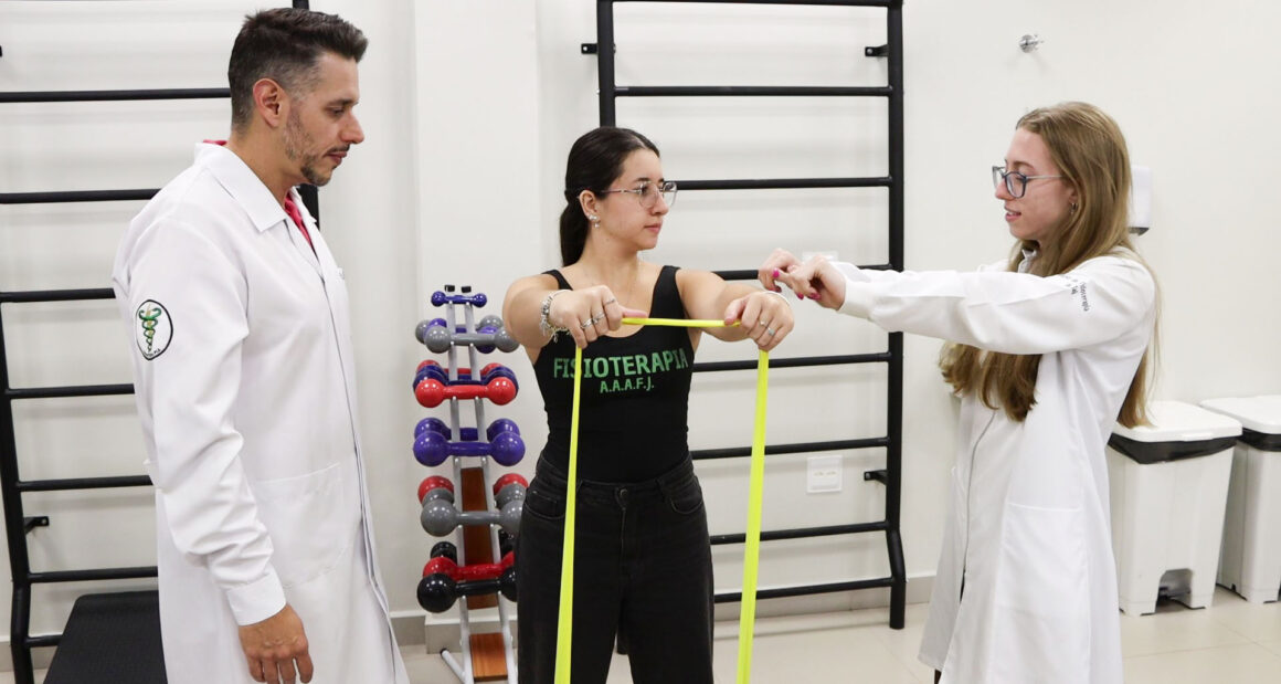 Professor e aluna do curso de Fisioterapia da Unoeste acompanham exercício com faixa elástica durante atividade prática em laboratório, com equipamentos de reabilitação ao fundo.