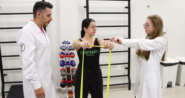 Professor e aluna do curso de Fisioterapia da Unoeste acompanham exercício com faixa elástica durante atividade prática em laboratório, com equipamentos de reabilitação ao fundo.