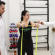 Professor e aluna do curso de Fisioterapia da Unoeste acompanham exercício com faixa elástica durante atividade prática em laboratório, com equipamentos de reabilitação ao fundo.