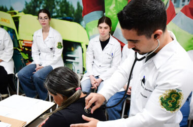 Estudante de Medicina utilizando estetoscópio para examinar paciente enquanto outros alunos acompanham a prática clínica em ambiente de aprendizado.