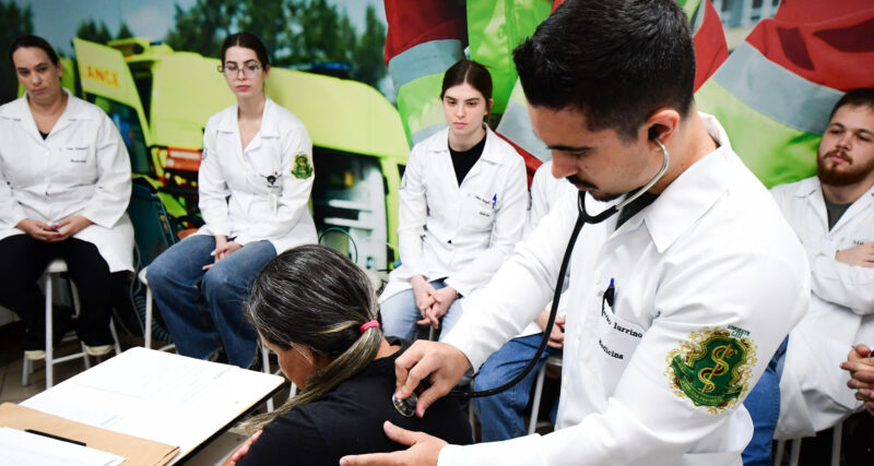 Estudante de Medicina utilizando estetoscópio para examinar paciente enquanto outros alunos acompanham a prática clínica em ambiente de aprendizado.
