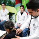 Estudante de Medicina utilizando estetoscópio para examinar paciente enquanto outros alunos acompanham a prática clínica em ambiente de aprendizado.