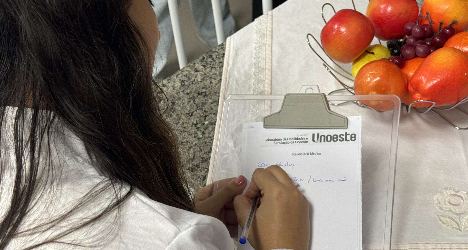 Estudante de Psicologia da Unoeste escreve anotações em um receituário durante atividade prática, ao lado de frutas utilizadas em dinâmica sobre alimentação e saúde.