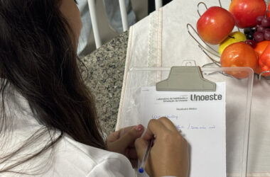 Estudante de Psicologia da Unoeste escreve anotações em um receituário durante atividade prática, ao lado de frutas utilizadas em dinâmica sobre alimentação e saúde.