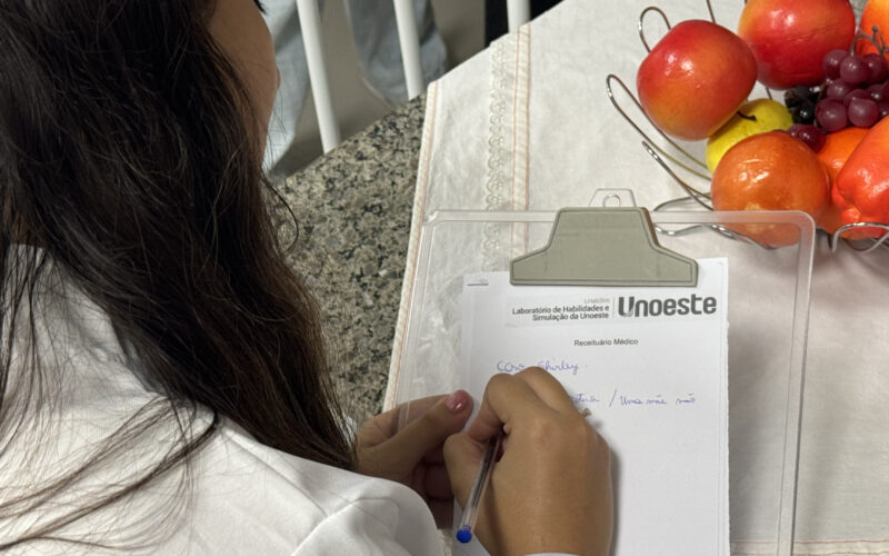 Estudante de Psicologia da Unoeste escreve anotações em um receituário durante atividade prática, ao lado de frutas utilizadas em dinâmica sobre alimentação e saúde.