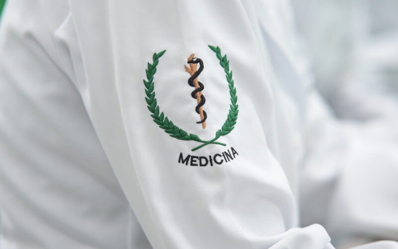Detalhe do jaleco de estudante de Medicina com bordado do símbolo da profissão na manga, representando a formação médica e a preparação para atuar na área da saúde.