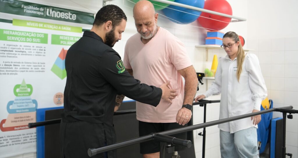 Estudantes de Fisioterapia da Unoeste acompanham um paciente durante atividade de reabilitação em barras paralelas. A prática ocorre em ambiente clínico da universidade e envolve avaliação, orientação e treinamento da mobilidade para recuperação da funcionalidade e da qualidade de vida.