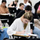 Estudantes universitários realizando prova em sala de aula, sentados em carteiras individuais e concentrados na resolução das questões de uma avaliação acadêmica.