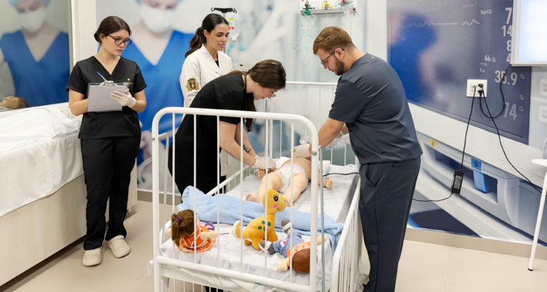 Estudantes e professores da área da saúde participam de uma atividade prática em um laboratório de simulação da Unoeste. Ao redor de um simulador pediátrico em um leito hospitalar, a equipe realiza procedimentos enquanto uma aluna registra informações em uma prancheta. O ambiente conta com equipamentos e recursos que reproduzem um cenário de atendimento hospitalar.