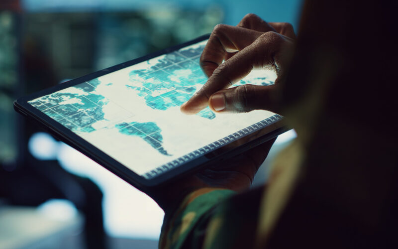 Close-up de uma pessoa utilizando os dedos para navegar em um mapa digital exibido em um tablet. A tela apresenta informações geográficas organizadas em áreas delimitadas, simulando uma análise realizada em um Sistema de Informação Geográfica (SIG), tecnologia utilizada para mapear territórios, analisar dados espaciais e apoiar a tomada de decisões em diferentes setores.