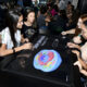 Estudantes participam de atividade colaborativa em uma mesa digital interativa durante aula prática, explorando um modelo tridimensional de célula em ambiente universitário.
