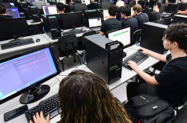 Estudantes utilizam computadores durante aula prática de programação em laboratório de informática, aprendendo linguagens de programação.