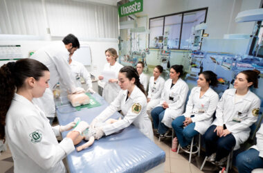 Estudantes de Medicina da Unoeste participam de aula prática em laboratório, com demonstração de procedimento em manequim pediátrico. Ao fundo, o ambiente conta com incubadoras e equipamentos que reproduzem uma unidade neonatal.