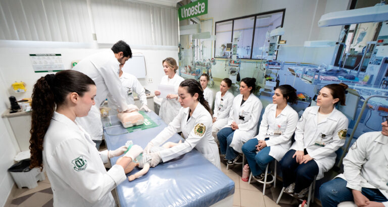 Estudantes de Medicina da Unoeste participam de aula prática em laboratório, com demonstração de procedimento em manequim pediátrico. Ao fundo, o ambiente conta com incubadoras e equipamentos que reproduzem uma unidade neonatal.