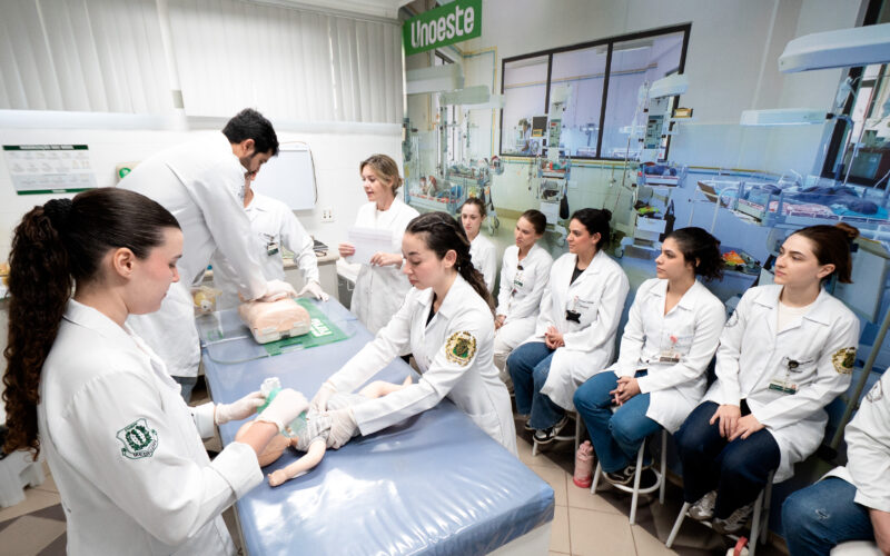 Estudantes de Medicina da Unoeste participam de aula prática em laboratório, com demonstração de procedimento em manequim pediátrico. Ao fundo, o ambiente conta com incubadoras e equipamentos que reproduzem uma unidade neonatal.