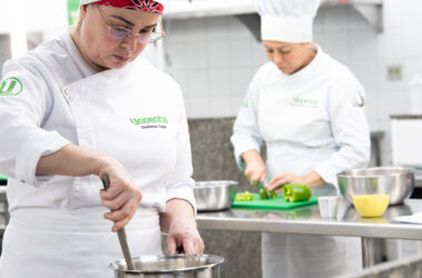 Duas alunas do curso de Gastronomia da Unoeste preparam alimentos durante aula prática em cozinha profissional, enquanto uma mexe uma panela e a outra corta legumes sobre uma bancada de inox.