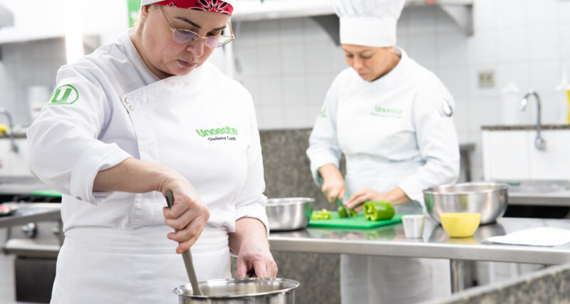 Duas alunas do curso de Gastronomia da Unoeste preparam alimentos durante aula prática em cozinha profissional, enquanto uma mexe uma panela e a outra corta legumes sobre uma bancada de inox.