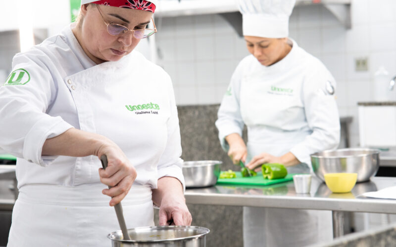 Duas alunas do curso de Gastronomia da Unoeste preparam alimentos durante aula prática em cozinha profissional, enquanto uma mexe uma panela e a outra corta legumes sobre uma bancada de inox.